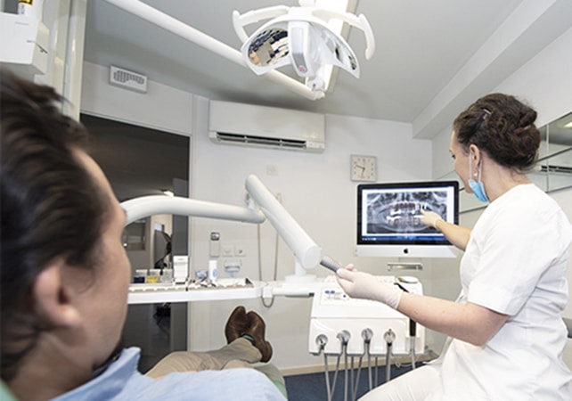 Zahnärztin berät Kunde für ein Zahnimplantat im Behandlungszimmer und zeigt Röntgenbild auf einem Screen